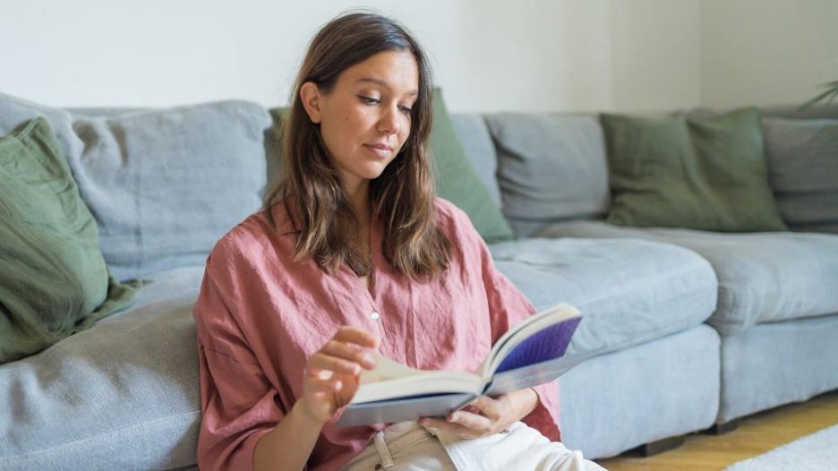 woman in a pink shirt reads a book while sitting up against a blue sofa